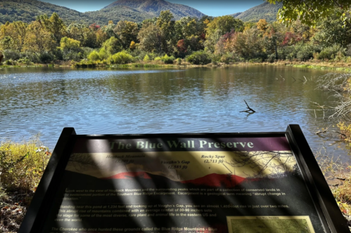 A scenic view of a lake surrounded by colorful autumn trees, with mountains in the background and an informational sign in the foreground.