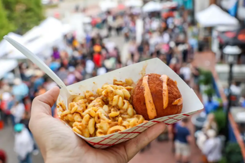 A hand holds a tray of pasta and a fried ball, with a busy outdoor food market in the background.
