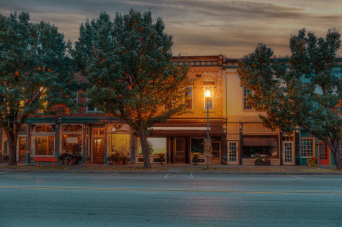 A quiet street scene at dusk, featuring charming storefronts and trees, illuminated by a streetlamp.