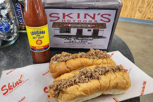 Two hot dogs topped with meat sauce, placed on a branded paper, alongside a bottle of hot sauce and a sign for Skin's Hotdogs.