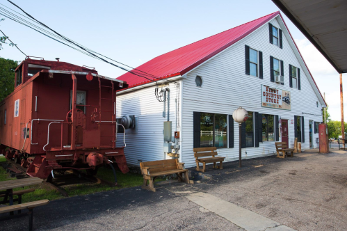 A red caboose next to a white country store with a red roof, featuring benches outside and a clear blue sky.