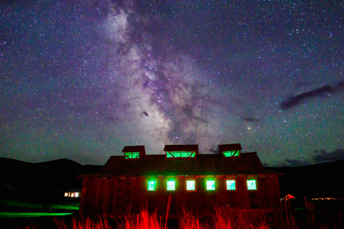 A starry night sky with the Milky Way above a rustic building illuminated by green lights.