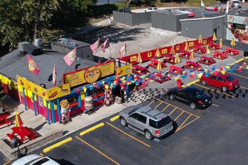 Aerial view of a colorful restaurant with outdoor seating, flags, and a parking lot filled with cars.