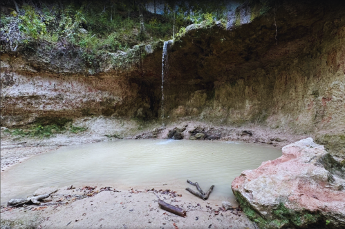 A serene natural pool surrounded by rocky cliffs and a small waterfall, with greenery in the background.
