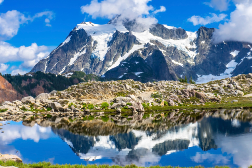 A stunning mountain landscape with snow-capped peaks reflected in a calm lake under a blue sky with fluffy clouds.
