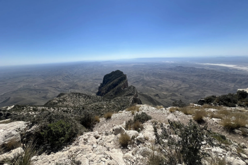 A panoramic view from a mountain peak overlooking a vast, arid landscape with distant mountains and clear blue sky.