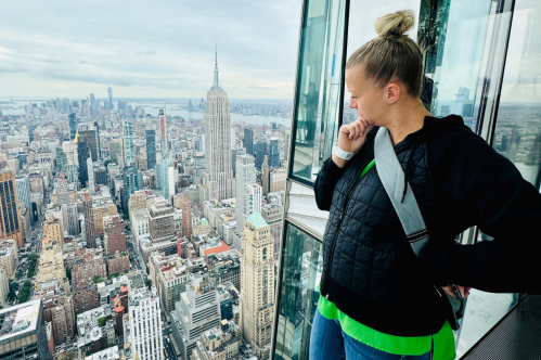 A woman gazes thoughtfully at the New York City skyline from a high vantage point, with the Empire State Building visible.