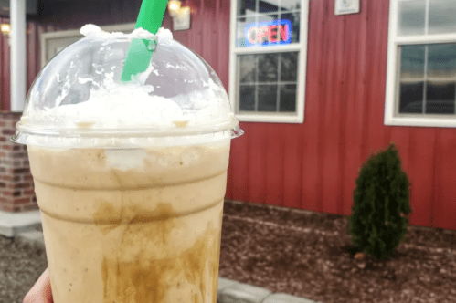 A hand holds a large iced drink topped with whipped cream, with a red building and "OPEN" sign in the background.