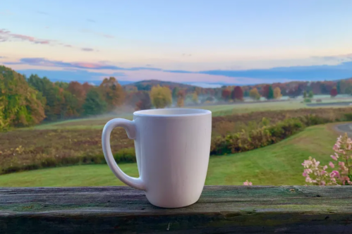 A white mug sits on a wooden railing, overlooking a scenic landscape with colorful autumn trees and a misty horizon.