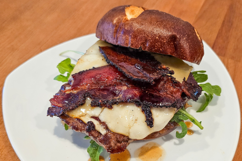 A pretzel bun sandwich with melted cheese, crispy bacon, and arugula on a white plate.