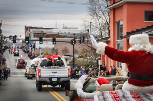 A festive parade scene with Santa waving from a convertible, surrounded by colorful floats and a cheering crowd.