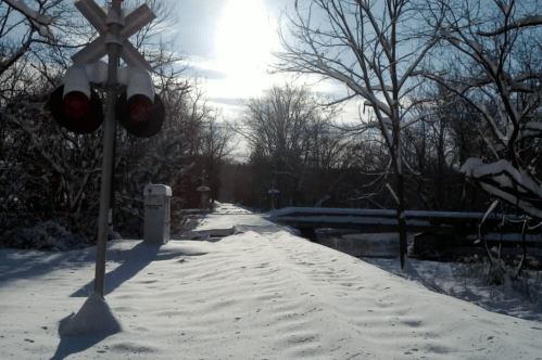A snowy landscape with a train crossing signal, trees in the background, and a bright sun shining in the sky.