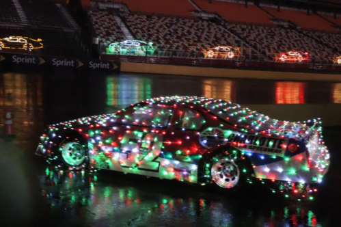 A brightly lit race car covered in colorful Christmas lights, parked on a wet racetrack at night.