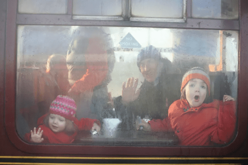 Two children and an adult wave from a foggy train window, smiling and wearing colorful winter coats.