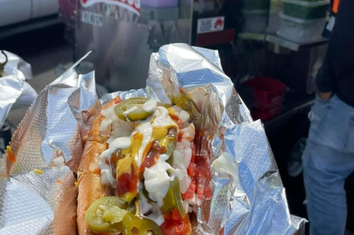 A close-up of a hot dog in foil, topped with onions, jalapeños, ketchup, and mustard, held in hand.