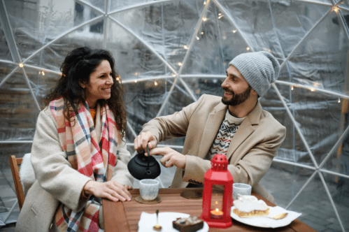 A couple enjoys a cozy moment in a transparent igloo, sharing drinks and smiles, with festive lights in the background.