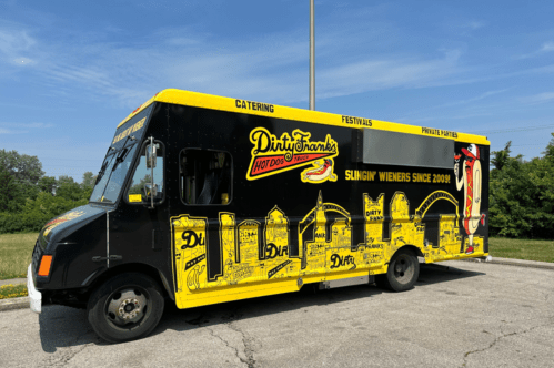 A colorful food truck with a yellow and black design, advertising Dirty Frank's hot dogs, parked in a grassy area.