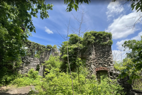 Ruins of a stone structure surrounded by lush greenery and trees under a bright blue sky with scattered clouds.