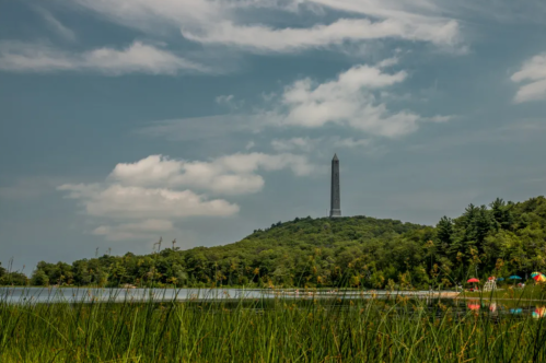 A tall monument stands on a hill, surrounded by trees and a calm lake under a cloudy sky.