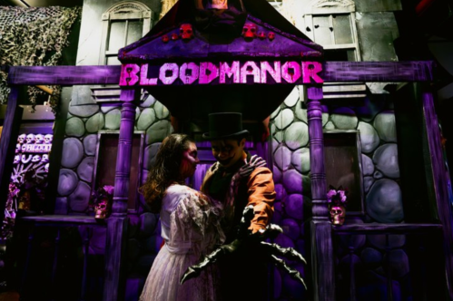 A spooky scene at "Blood Manor" featuring a couple in costume, with eerie decorations and purple lighting.