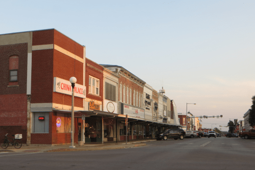 A quiet street scene featuring storefronts, including a Chinese restaurant, under a soft evening sky.