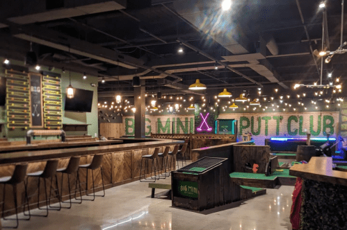 Interior of a mini putt club featuring a bar, colorful lighting, and a vibrant putting area with decorative elements.