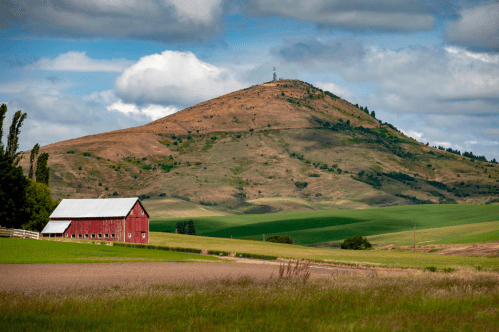A red barn sits in a green field with a hill in the background under a partly cloudy sky.