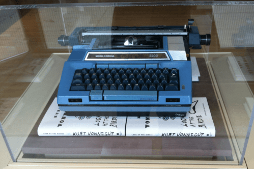 A blue Smith-Corona 2200 typewriter displayed in a glass case, with Kurt Vonnegut's book "Look at the Birdie" underneath.