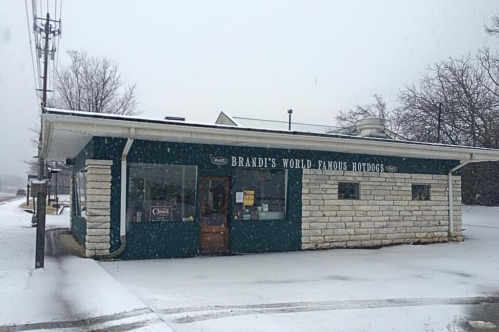 A small, snow-covered building with a sign reading "Brandt's World Famous Hotdogs" on a winter day.