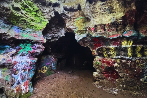 A colorful cave interior with graffiti on the rocky walls and a dark entrance leading deeper into the cave.