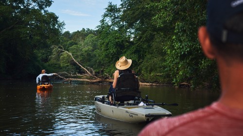 Two people kayaking on a calm river surrounded by lush greenery, one wearing a straw hat.