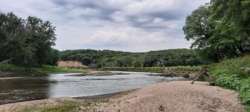 A serene riverbank scene with sandy shores, lush greenery, and overcast skies in the background.