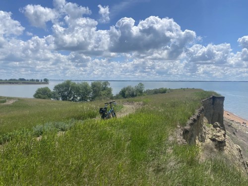 A bicycle rests on a grassy cliff overlooking a calm lake under a partly cloudy sky.