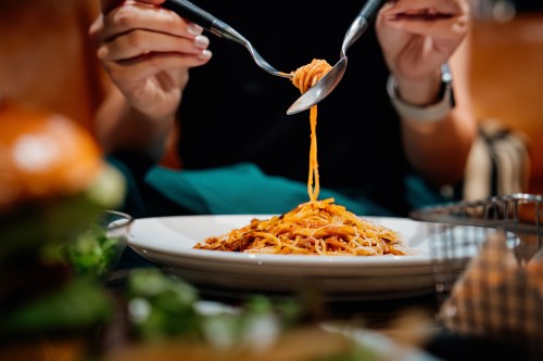 A person using forks to twirl spaghetti on a plate, with a blurred burger and salad in the background.