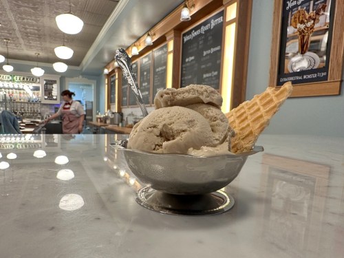 A silver bowl filled with three scoops of ice cream and a waffle cone, set on a marble counter in an ice cream shop.