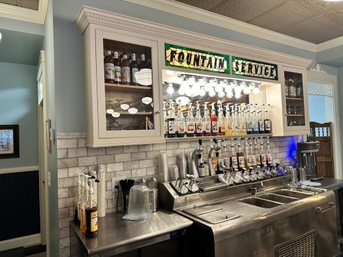 A vintage-style soda fountain with various syrup bottles and glassware displayed in a well-lit café setting.