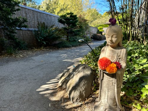 A serene stone statue of a Buddha surrounded by greenery and flowers along a peaceful garden path.