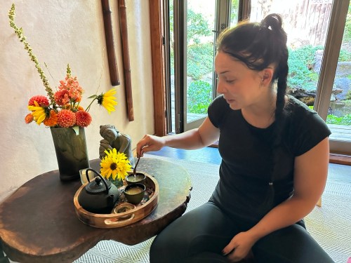 A woman in a black shirt prepares tea at a wooden table adorned with flowers and a decorative statue.