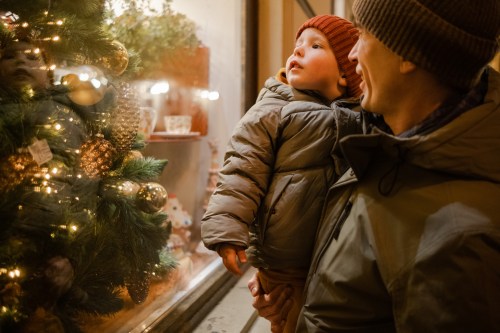 A parent holds a child, both gazing at a festive window display adorned with Christmas decorations.