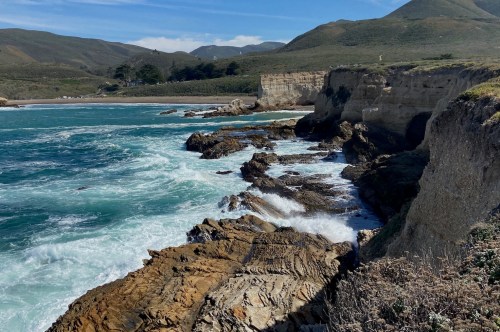 A rocky coastline with crashing waves, cliffs, and green hills under a clear blue sky.