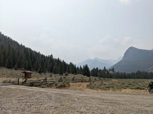 A gravel road leads to a wooden structure, surrounded by trees and mountains under a hazy sky.