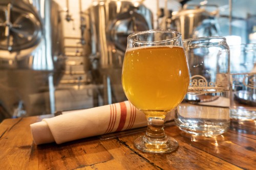 A glass of golden beer sits on a wooden table, with water glasses and brewing equipment in the background.