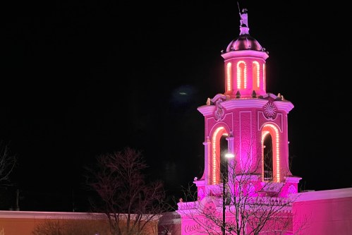 A brightly lit tower with pink lights at night, featuring decorative elements and a statue on top.
