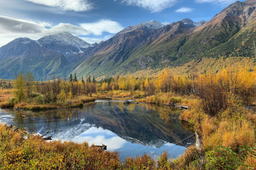 A serene landscape featuring a calm pond surrounded by autumn foliage and mountains under a blue sky with clouds.