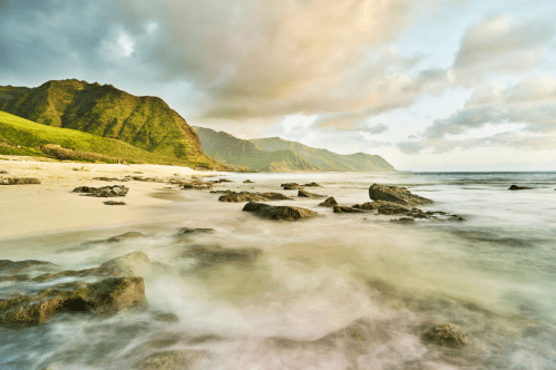 A serene beach scene with smooth waves, rocky shoreline, and lush green mountains under a cloudy sky.