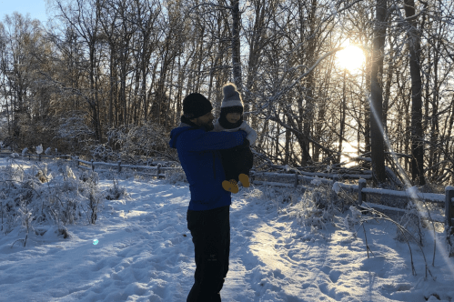 A parent holds a child in a snowy landscape, with trees and a setting sun in the background.