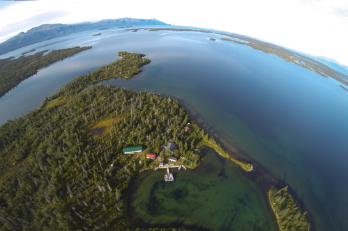Aerial view of a serene lake surrounded by lush forests and small islands, with a cabin and dock visible on the shore.