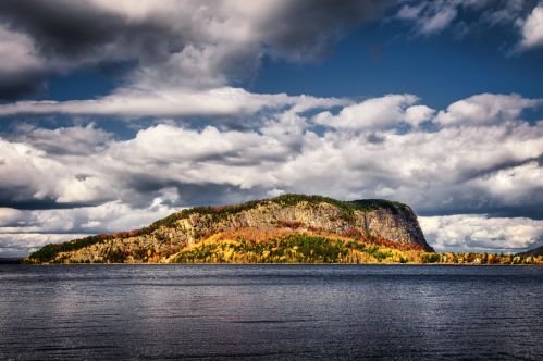 A scenic view of a rocky island surrounded by water, with colorful autumn foliage and dramatic clouds overhead.