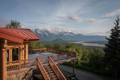 A scenic view from a wooden deck overlooking mountains and a river, with a stone house in the foreground.