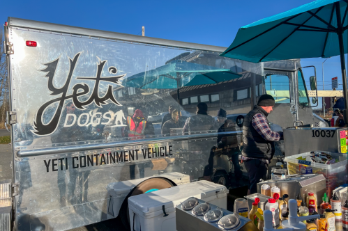 A food truck named "Yeti Dogs" with a shiny exterior, featuring a vendor preparing food under blue umbrellas.